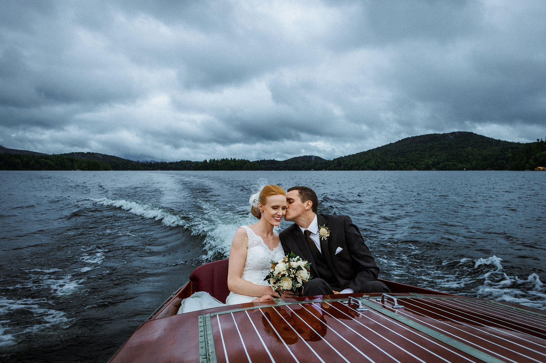 Lakeside wedding portrait of a bride and groom kissing aboard a classic wooden boat